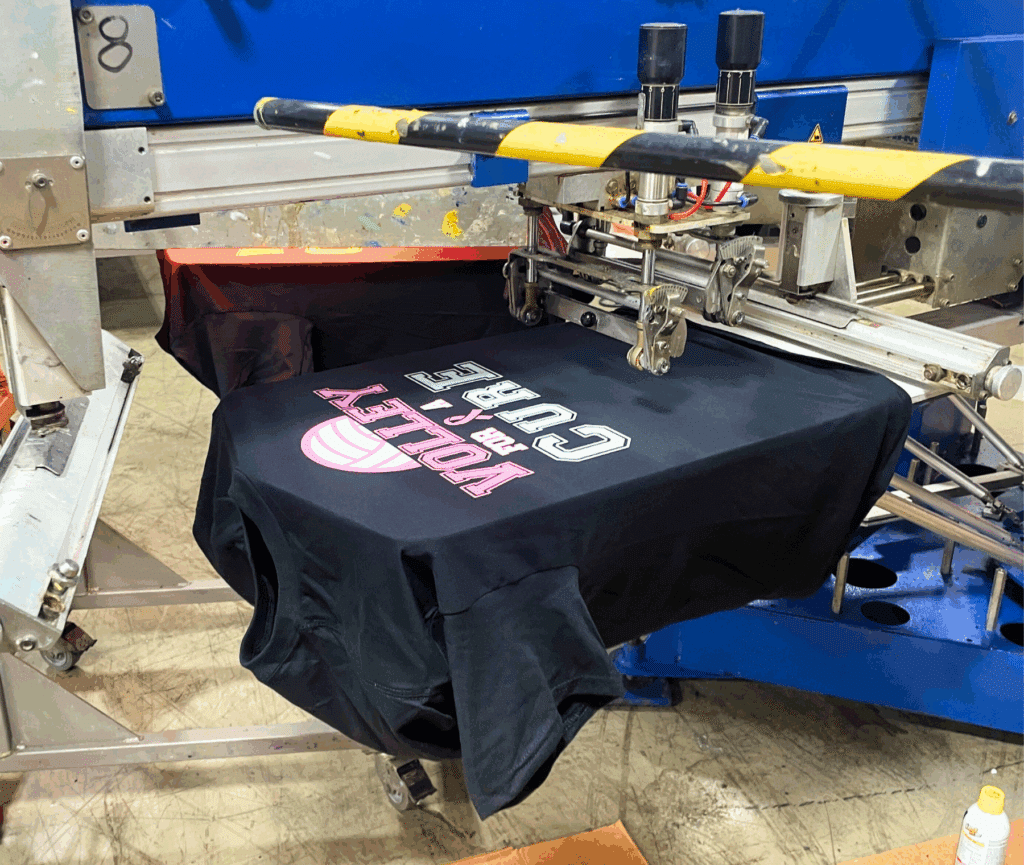 Industrial screen printing process applying a custom ‘Volley for a Cure’ design onto a black T-shirt.