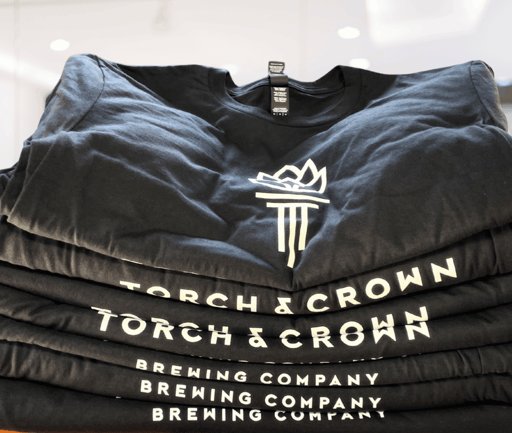 Stack of black Torch & Crown Brewing Company custom printed T-shirts with white logo design.
