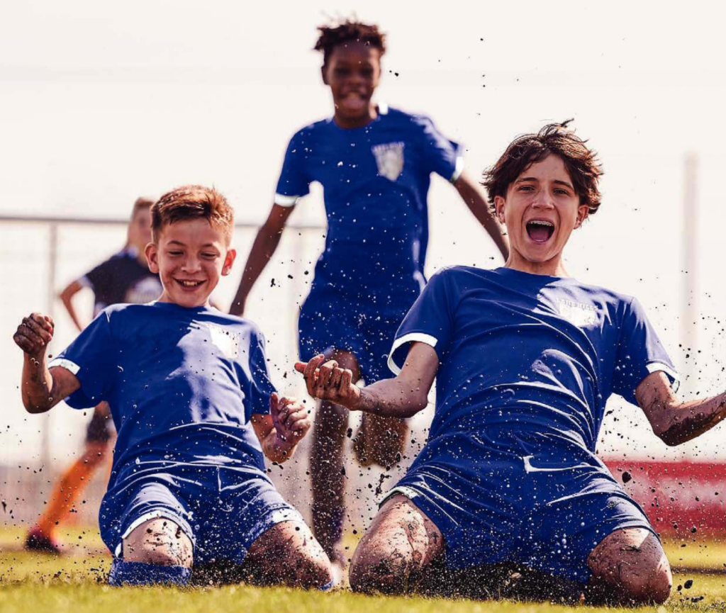 Youth soccer players in blue uniforms sliding on grass and celebrating during a game.